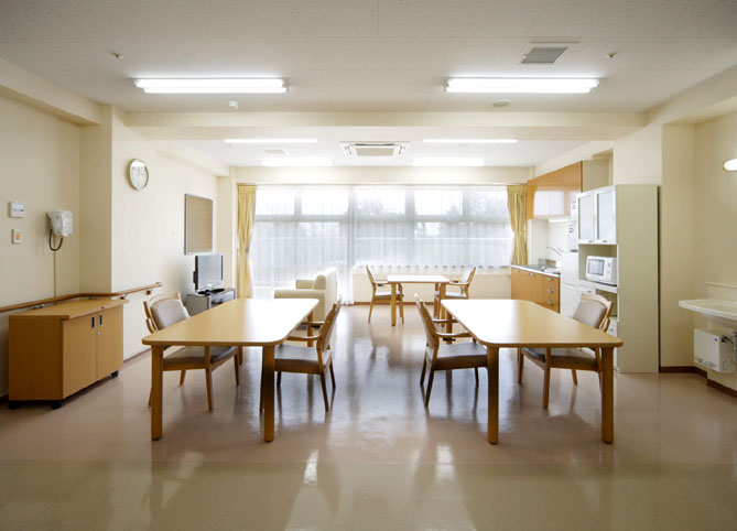 介護施設の食堂。木製テーブルと椅子が配置され、明るく清潔な共有スペース。