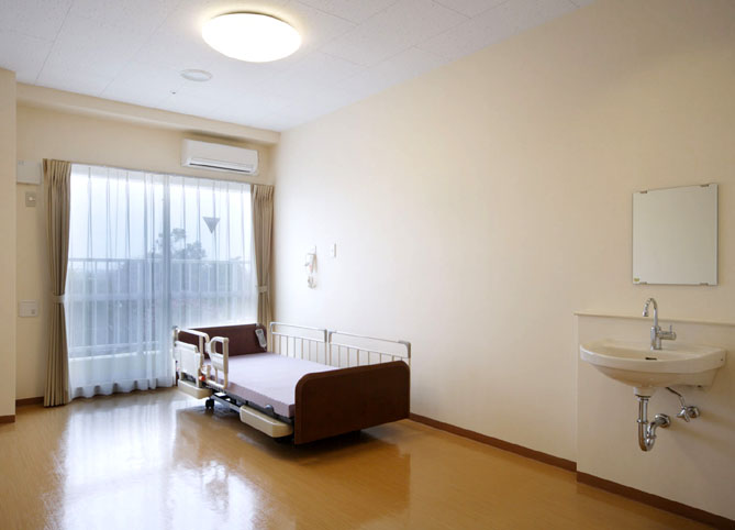 介護ベッド、洗面台、窓を備えた個室。明るく清潔な空間。